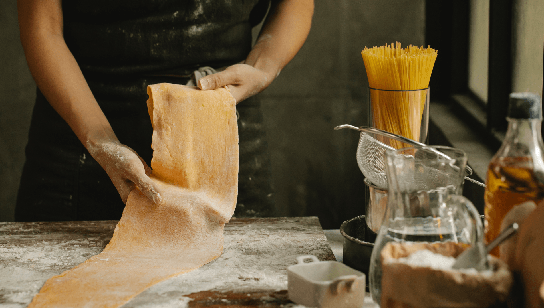 Massa Fresca Italiana: Da Escolha da Farinha ao Corte Perfeito 1 Mãos amassando massa fresca italiana em uma bancada enfarinhada, com rolo e massa aberta ao fundo