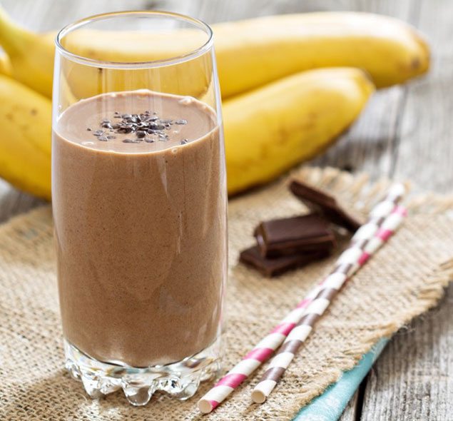 Shake de Banana, Aveia e Cacau com Proteína 1 Copo alto com shake roxo-esverdeado, canudo, fatias de banana e aveia ao lado, fundo de madeira rústica